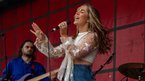 MACY MACY singing on stage holding a microphone.  Her bass player, a man, stands behind her.